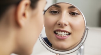 Primer plano de una mujer joven examinando el esmalte de sus dientes en un espejo de mano, que refleja su boca, nariz y ojos.