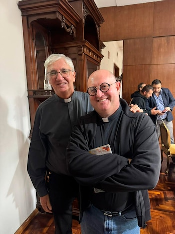Dos sacerdotes sonríen a cámara: uno calvo con gafas redondas, chaqueta negra y jeans, y otro con pelo gris y gafas, camisa clerical. Fondo de madera y mueble