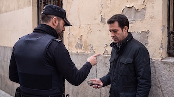 Un policía de uniforme apunta con el dedo a un hombre con abrigo que sostiene un teléfono móvil frente a una pared deteriorada.
