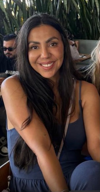 Una mujer de cabello oscuro y sonrisa posa en primer plano. Lleva un top azul oscuro. Al fondo hay vegetación y un hombre con gafas de sol