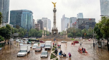 Vista aérea de una calle inundada con varios coches, autobuses y personas transitando; algunos usan balsas. Al fondo se observa un monumento y edificios.