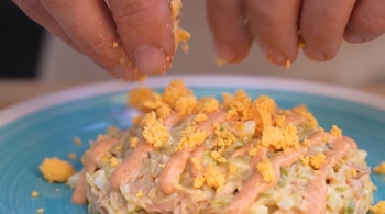 Dos manos esparcen trozos pequeños de yema de huevo cocido sobre una ensalada mezclada con salsa rosa y otros ingredientes en un plato azul.
