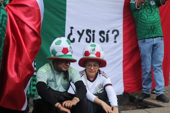 Dos personas con sombreros de fútbol y camisetas deportivas. Detrás, una bandera de México con el texto "¿Y si sí?"