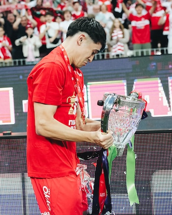 Un hombre joven vestido con camiseta roja sostiene un trofeo de cristal y metal roto, con el vidrio fragmentado. De fondo, una pantalla LED y una multitud
