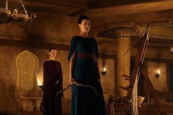 Sala con dos mujeres, columnas y antorchas. La mujer delantera, con vestido azul y collar, sujeta un arco. La mujer trasera usa vestido granate