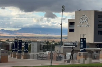 Vista exterior de un edificio del estadio de Utah State University con el logo del toro, montañas y cielo nublado. Se ve una estatua