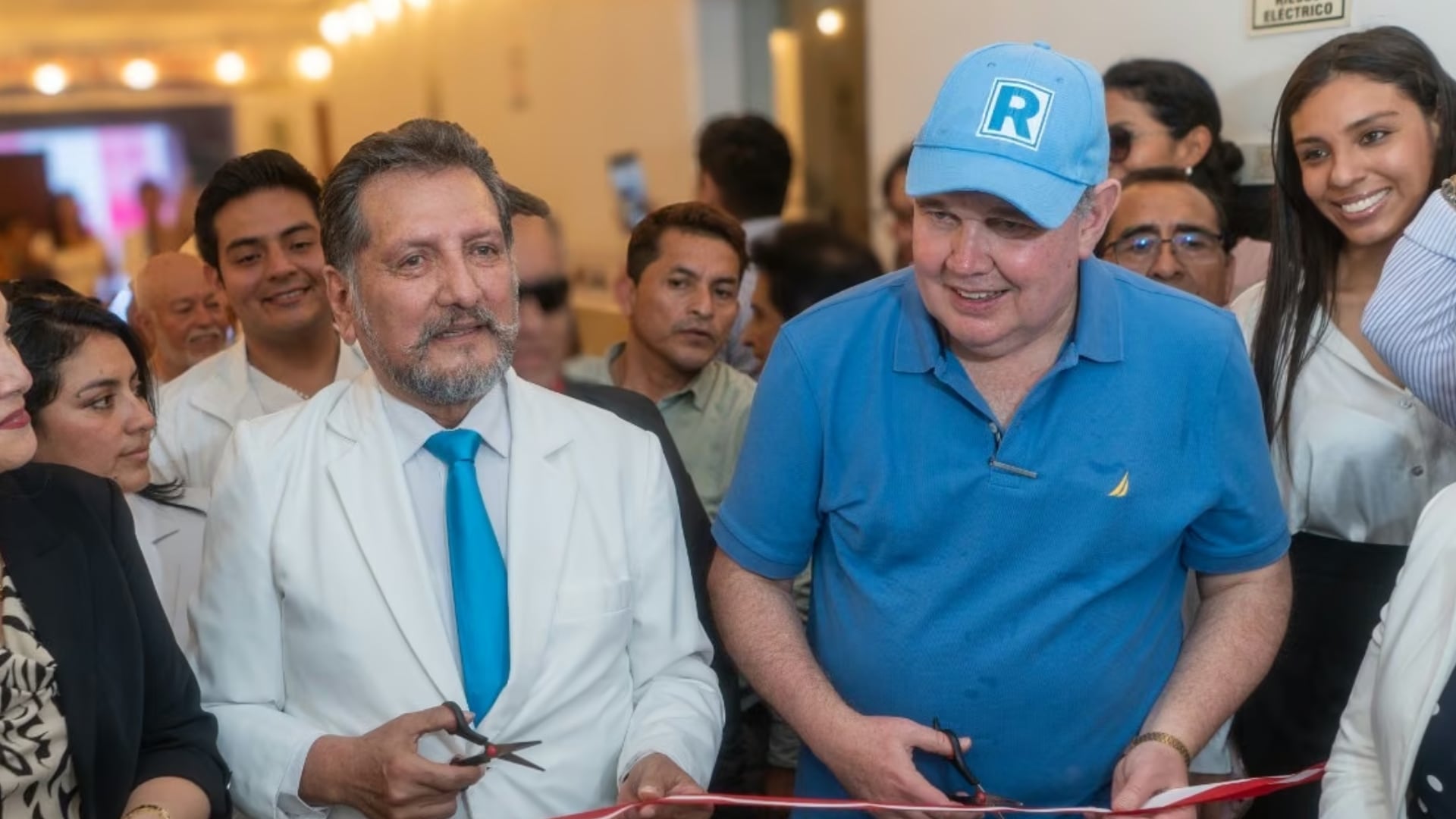 Rafael López Aliaga, con gorra azul y polo del mismo color, y el exfuncionario Luis Rubio, con saco blanco y corbata celeste, participan en el corte de una cinta roja durante un evento rodeados de personas.