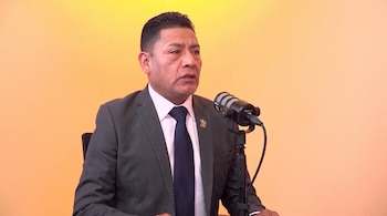 Un hombre vestido con traje gris y corbata azul habla frente a un micrófono negro sobre un fondo naranja claro.