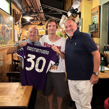 Tres hombres de pie en un restaurante sostienen una camiseta de fútbol morada con el número 30 y el apellido Mastantuono