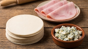 Una pila de tapas de empanadas redondas, un plato con lonchas de jamón cocido dobladas y un cuenco con queso roquefort desmenuzado sobre una mesa de madera.
