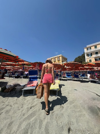 Mujer de espaldas camina por una playa de arena con sombrillas y reposeras. Viste bikini rojo a cuadros, falda, sombrero de paja y lleva un bolso de mimbre