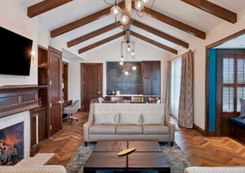 Sala de estar de hotel con techo de vigas de madera, chimenea encendida, sofá beige, mesa de centro oscura y un área de comedor al fondo