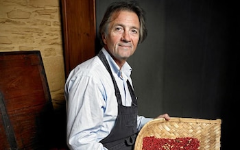 Hombre de mediana edad, John Powell, sonríe con camisa azul claro y delantal oscuro, sosteniendo una canasta de mimbre llena de pimienta rosa y anís estrellado