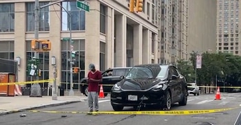 Calle acordonada con cinta amarilla, un automóvil deportivo negro con daños frontales, un hombre con gorra y teléfono, conos naranjas, semáforos, edificios