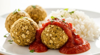 Albóndigas de lenteja y avena doradas en un plato blanco, cubiertas con salsa de tomate casera y acompañadas de arroz blanco. Fondo luminoso.