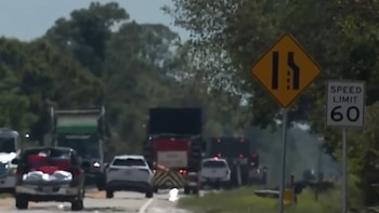 Vista frontal de una carretera con tráfico de vehículos y camiones, con una señal amarilla de estrechamiento de carril y una de límite de velocidad de 60
