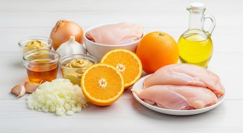 Pechugas de pollo, naranjas enteras y cortadas, cebolla entera y picada, ajos, mostaza, miel y aceite en recipientes sobre una mesa de madera clara.
