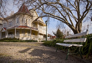 Casa de dos pisos con tejado rojo y balcones. Camino de grava, banco de madera antiguo, farola, árbol sin hojas y vegetación