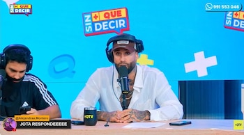 Jota Benz sentado en un estudio de televisión, con una gorra, auriculares y camisa blanca, hablando a un micrófono. Un hombre al lado lo mira
