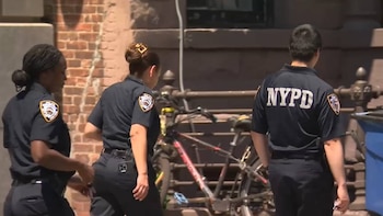 Vista trasera de tres oficiales del NYPD con uniformes oscuros caminando por una calle con edificios de ladrillo y una bicicleta estacionada