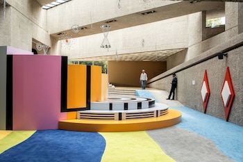 Interior de museo con paredes de hormigón, dos personas, una instalación artística de paneles rosas y naranjas, una plataforma circular escalonada y alfombras de colores