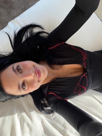 Una mujer de cabello negro recostada en una cama viste una prenda deportiva negra con costuras rojas