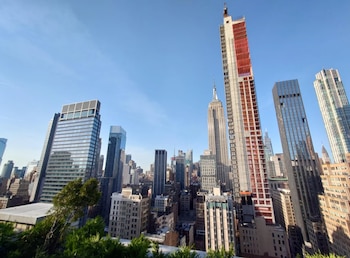 Vista aérea de un paisaje urbano con un rascacielos en construcción con malla roja y el Empire State Building, entre otros edificios altos, bajo un cielo azul claro
