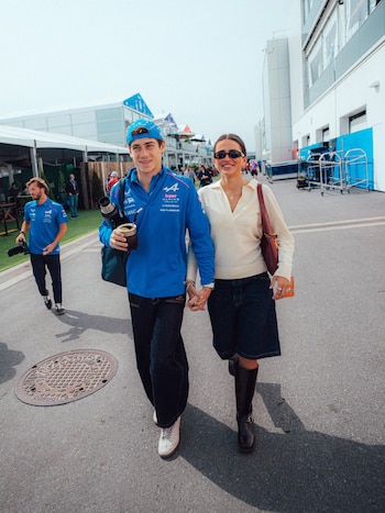 Franco Colapinto y Maia Reficco caminan al aire libre, tomados de la mano. Él lleva una chaqueta azul Alpine y un mate; ella, un suéter crema y falda denim