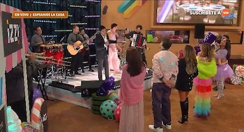 Un grupo de músicos toca instrumentos en un escenario mientras varias personas con prendas coloridas los observan en un estudio de televisión