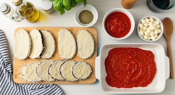 Rodajas de berenjena empanadas en tabla, salsa de tomate, bolas de mozzarella, orégano, hojas de albahaca, aceite de oliva, salero, bandeja y paño.