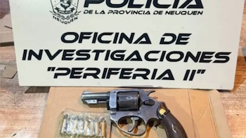 Un revólver gris oscuro y un paquete de balas sobre una superficie marrón, frente a un cartel blanco de la Policía de Neuquén