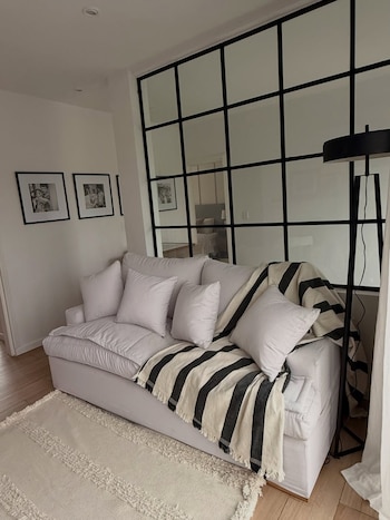 Sofá blanco con almohadones y manta a rayas blancas y negras en una sala de estar. Hay una alfombra clara, un piso de madera, y una pared divisoria de vidrio con marcos negros