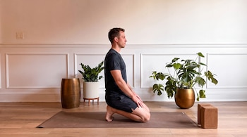 Hombre arrodillado de perfil en colchoneta de yoga, manos sobre rodillas. Fondo con pared blanca, dos plantas, un taburete y bloques de yoga.