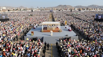 Un escenario con sacerdotes y el papa frente a una multitud con banderas en una explanada con pantallas gigantes y cerros al fondo.