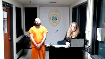 Hombre calvo y barbudo en mono naranja de prisión de pie en una sala de tribunal junto a una mujer con cabello largo sentada frente a un portátil y un micrófono