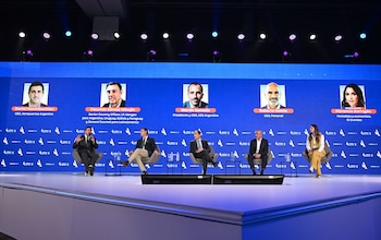 Cinco panelistas en un escenario moderno con iluminación. Una pantalla azul muestra las fotos, nombres y cargos de los oradores del AmCham Summit