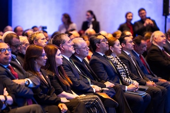 Filas de personas sentadas con atuendo formal, incluyendo trajes y camisas, miran hacia el frente en una sala con iluminación azul de fondo