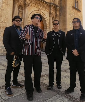 Cuatro hombres con chaquetas oscuras y gafas de sol estan de pie sobre un pavimento frente a una portada de piedra con arcos
