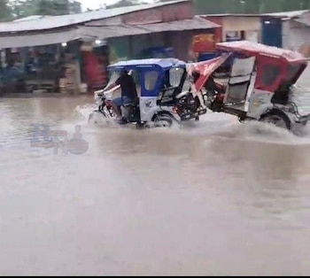 Dos mototaxis, uno azul y otro rojo, se abren paso en una calle de Pucallpa con un alto nivel de agua turbia, con edificaciones al fondo