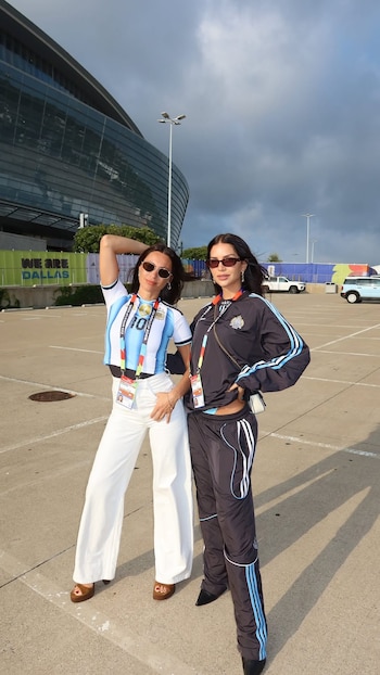 Dos mujeres con acreditaciones y gafas de sol posan frente a un estadio con un cartel que indica "WE ARE DALLAS". Una viste camiseta argentina, la otra un conjunto deportivo
