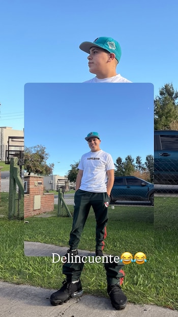 Un joven con gorra turquesa y blanca, camiseta blanca y pantalón oscuro con franjas rojas, posa sonriente al aire libre sobre un sendero de cemento