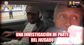 Un hombre con gorra y chaqueta se sienta en el asiento trasero de un coche, mirando hacia adelante. Un hombre mayor se ve a su lado. Se ve a un guardia fuera del auto