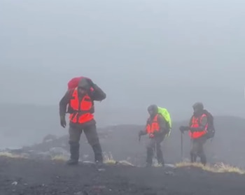 Tres excursionistas con chalecos reflectantes y mochilas caminan por un terreno rocoso y oscuro envuelto en una densa niebla