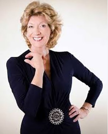 Retrato de Felicity Lott, una mujer de cabello rubio rizado, vestida de azul oscuro con un broche brillante, sonriendo levemente y tocando su barbilla