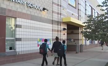 Tres niños con mochilas caminan tomados de la mano hacia la entrada de una escuela primaria que tiene un cartel de bienvenida
