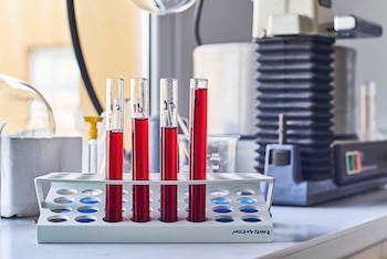 Un soporte blanco con cuatro tubos de ensayo llenos de líquido rojo. Los tubos tienen números escritos. Hay equipo de laboratorio en el fondo