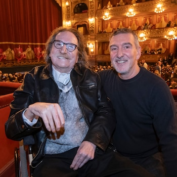 Charly García, con gafas y bigote, sonríe en una silla de ruedas al lado de un hombre canoso que lo abraza, ambos en el interior del Teatro Colón