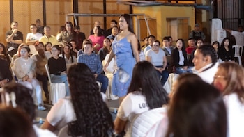 Una mujer con un vestido azul desfila en una pasarela al aire libre, observada por un público sentado en sillas blancas y personas de pie al fondo