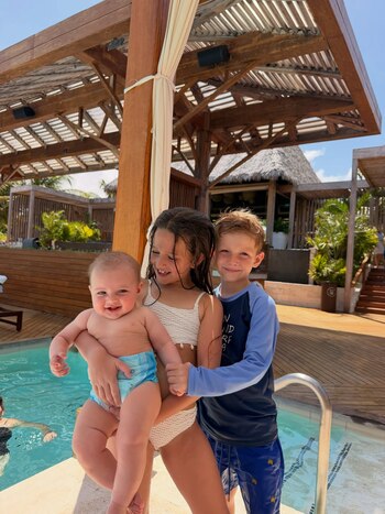 Tres niños sonríen junto a una piscina al aire libre; una niña sostiene a un bebé, mientras un niño rubio se para a su lado. El fondo tiene una estructura de madera y techo de paja