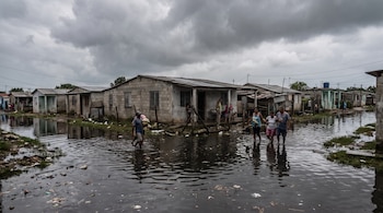 Varias personas caminan por una calle inundada frente a casas de bloques de hormigón bajo un cielo gris.